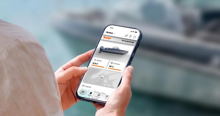 In the foreground, a smartphone in a person’s hands. The person’s upper arm is also visible. The Buster App is displayed on the screen. A Buster boat is visible in the blurred background.