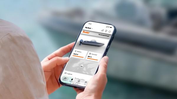 In the foreground, a smartphone in a person’s hands. The person’s upper arm is also visible. The Buster App is displayed on the screen. A Buster boat is visible in the blurred background.